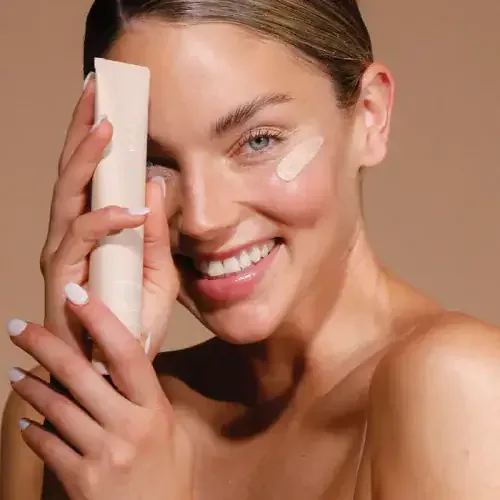 Smiling model holding an Airyday Dreamscreen SPF50+ tube beside her eye, warm tan backdrop