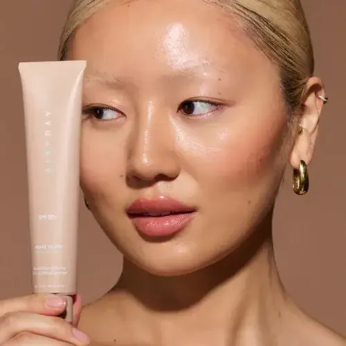 Model holding an Airyday Pearl Glow SPF50+ tube against her cheek, gold hoop earring